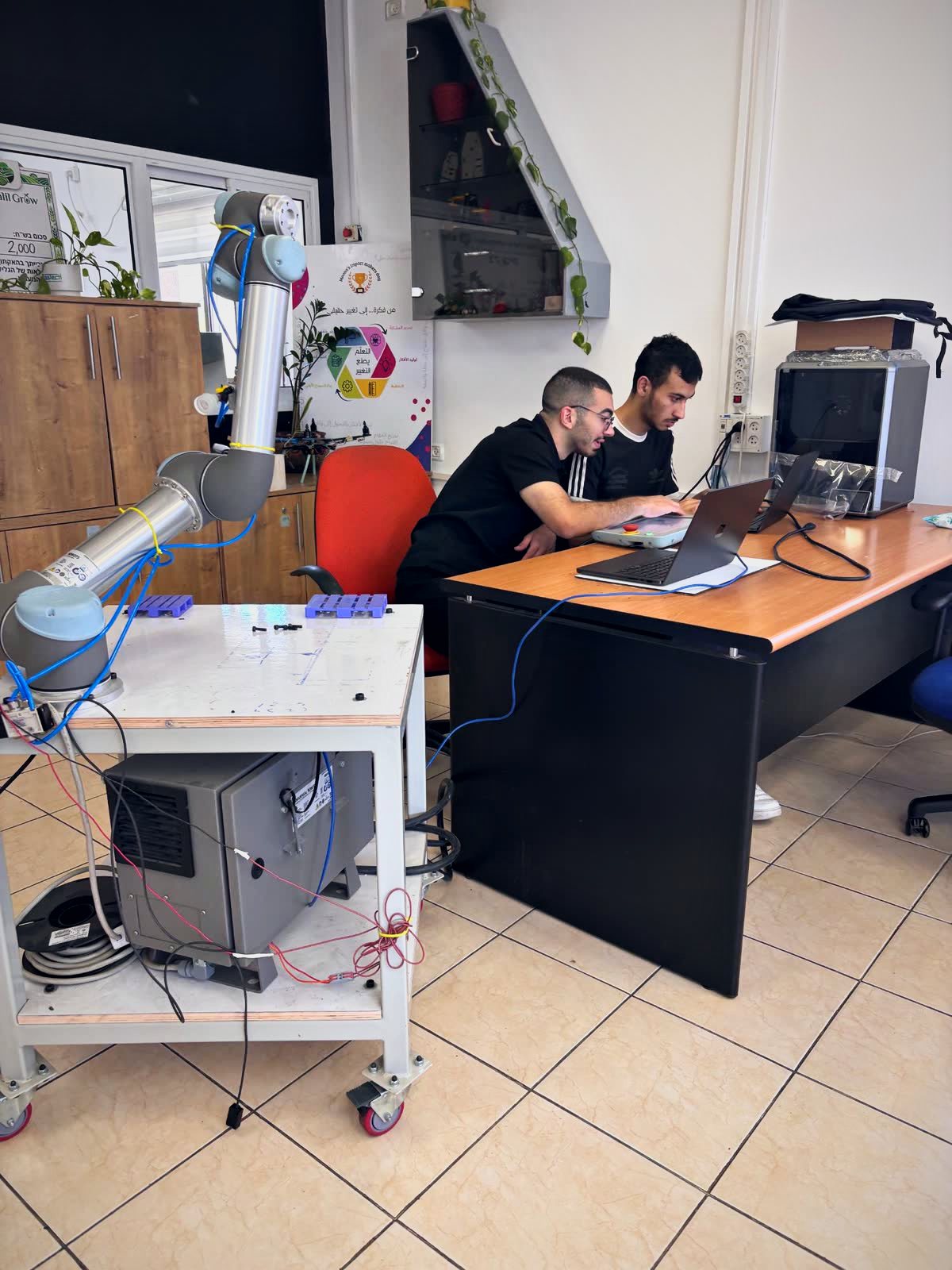 Two team members programming a UR5e cobot from a desk with laptops and the teach pendant