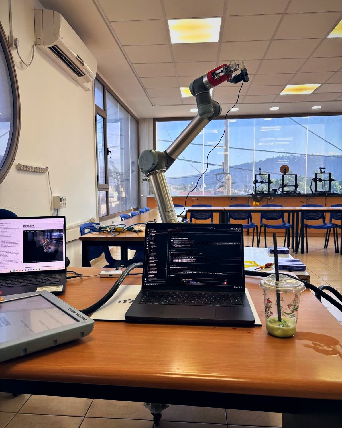 UR5 robot arm with a gripper and wrist camera above a desk with laptops running code, one showing a scan of the cell, in a lab with 3D printers